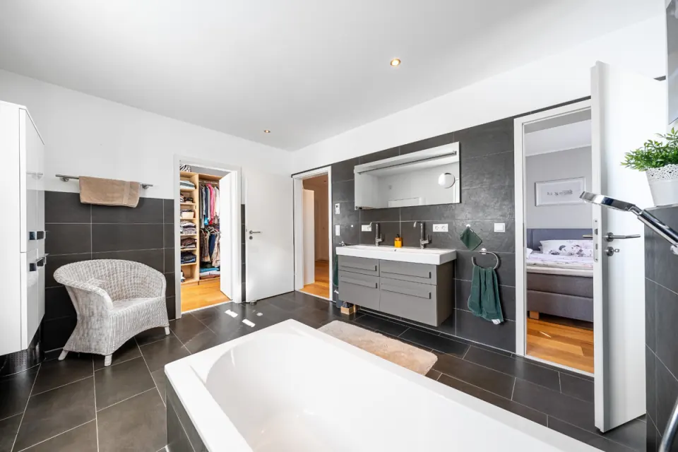 Badezimmer mit dunklen Großformatfliesen, Doppelwaschtisch, Badewanne und Blick ins Schlafzimmer