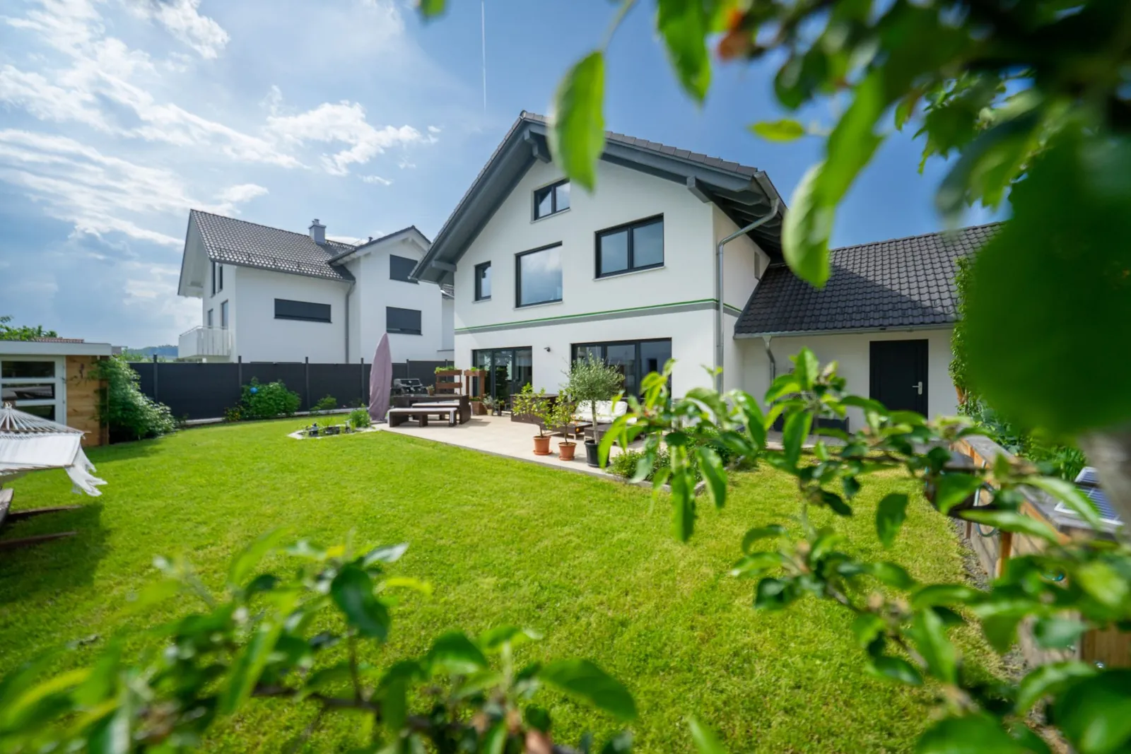 Weißes Einfamilienhaus mit Satteldach, großer Rasenfläche und Terrasse, durch Blätter im Vordergrund fotografiert
