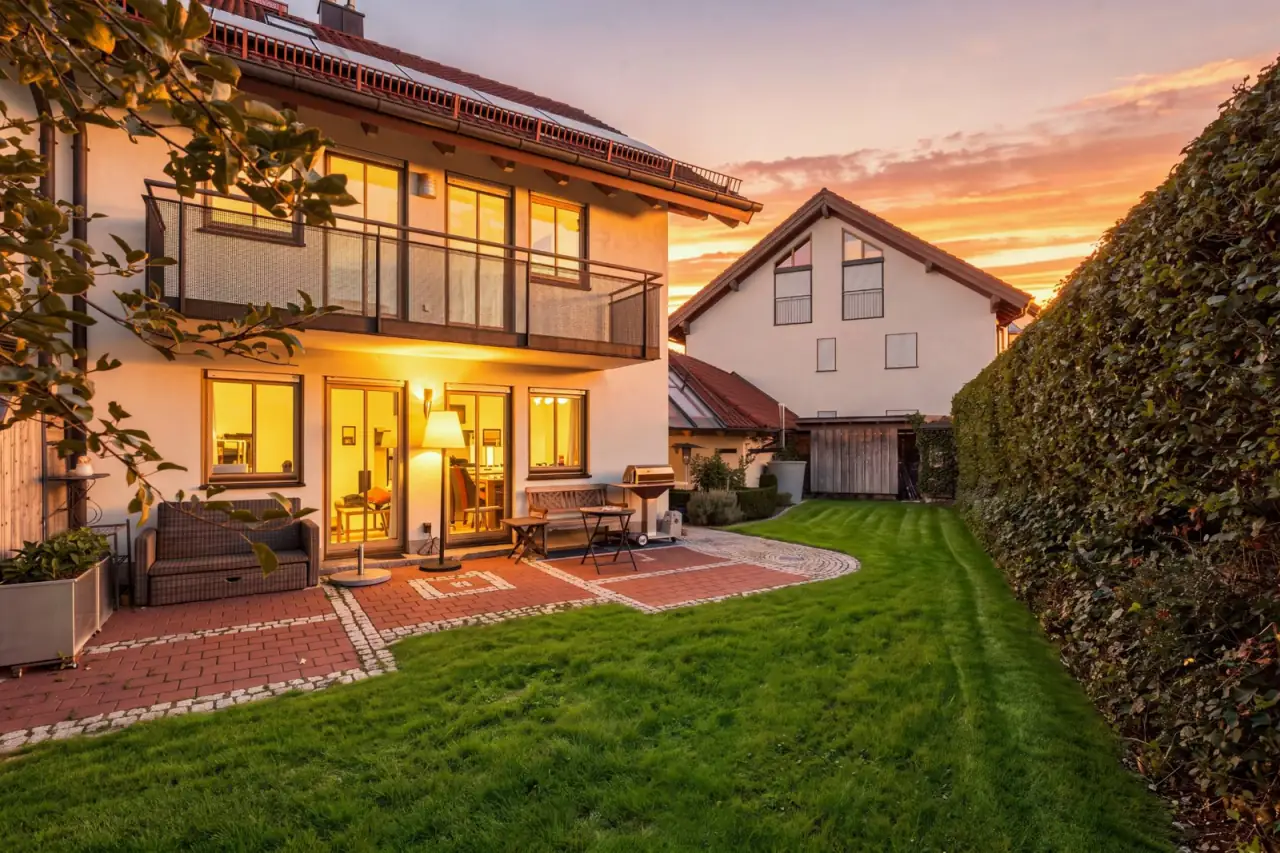 Doppelhaushälfte mit Balkon, beleuchteten Fenstern, Terrasse und Rasen im Abendlicht